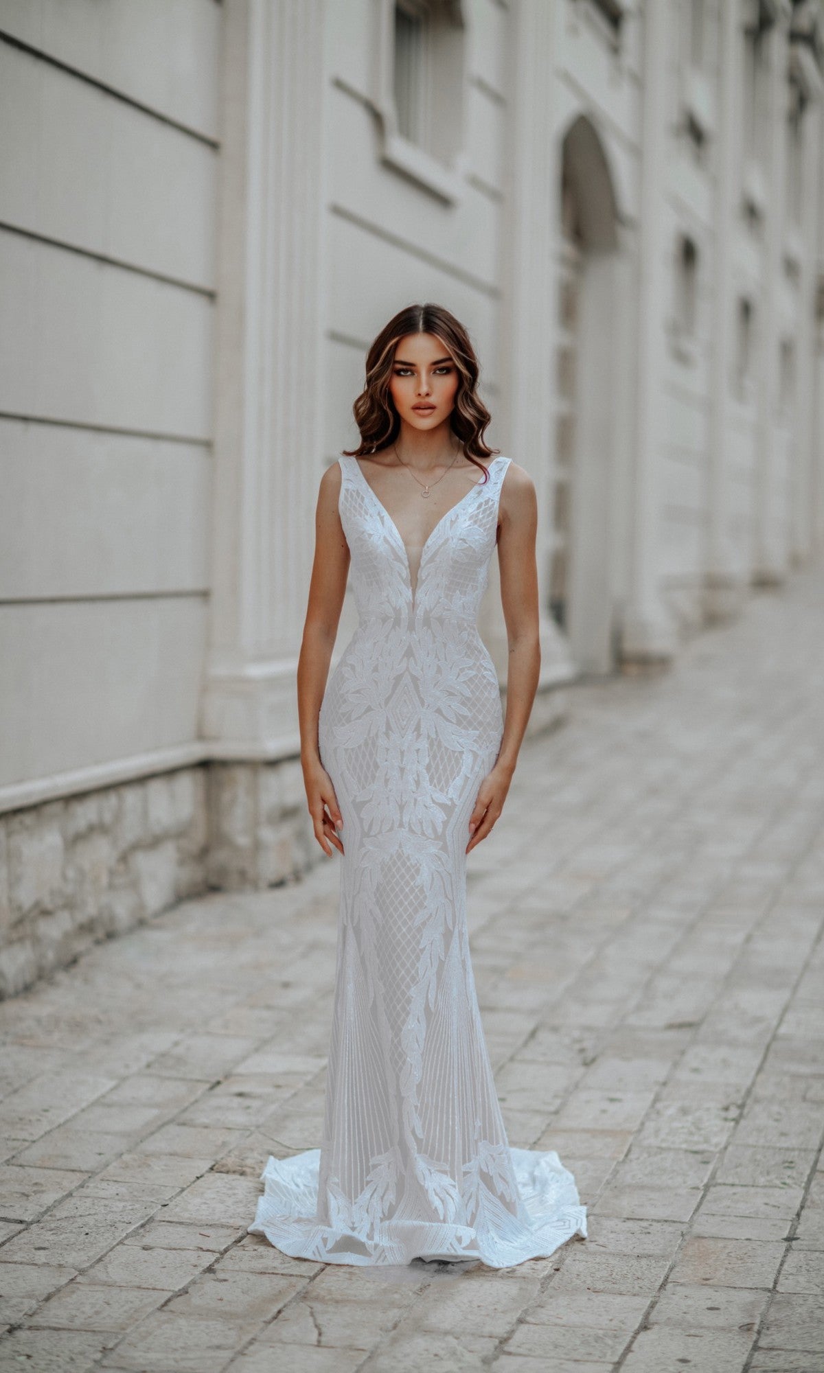A woman in the Classic Long White Formal Dress: Tina Holly BB052, a sleeveless, floor-length gown with lace detailing, stands on a stone walkway beside a light-colored building.